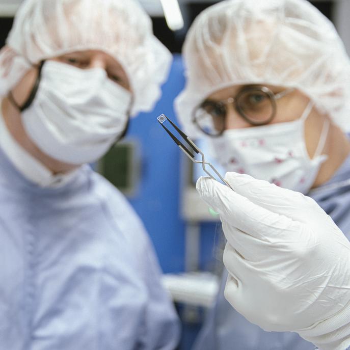 Photo with two persons in cleanroom suits looking at a miniaturized implant held by tweezers.