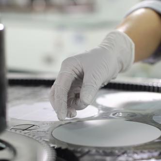 A white-gloved hand hovers over a thin white disk during waveguide production