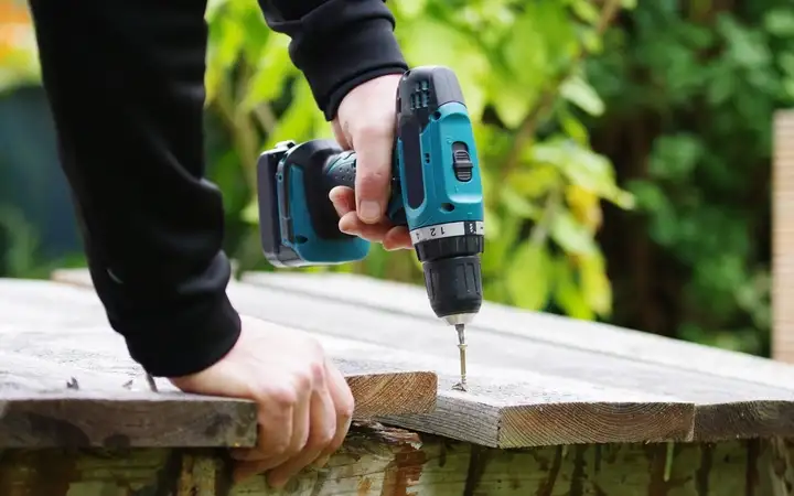 A person uses a cordless drill to fasten a board on a deck.