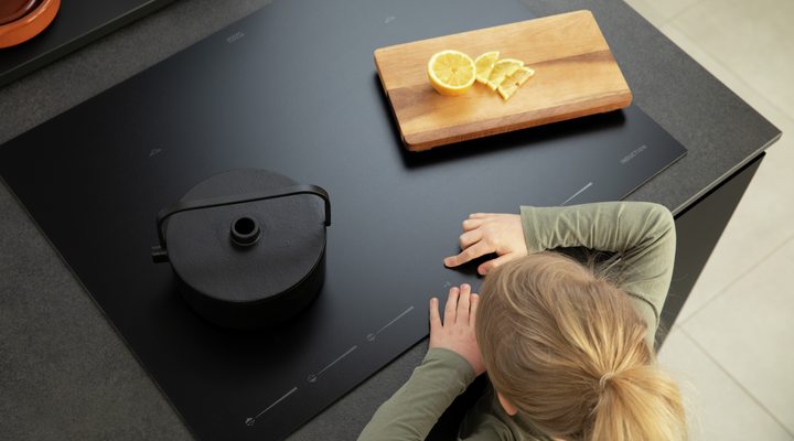 Top view of black glass cooktop with pot, cutting board with lemon slices, and person nearby
