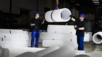 Two workers standing between large industrial pipes inside a quartz glass production facility.