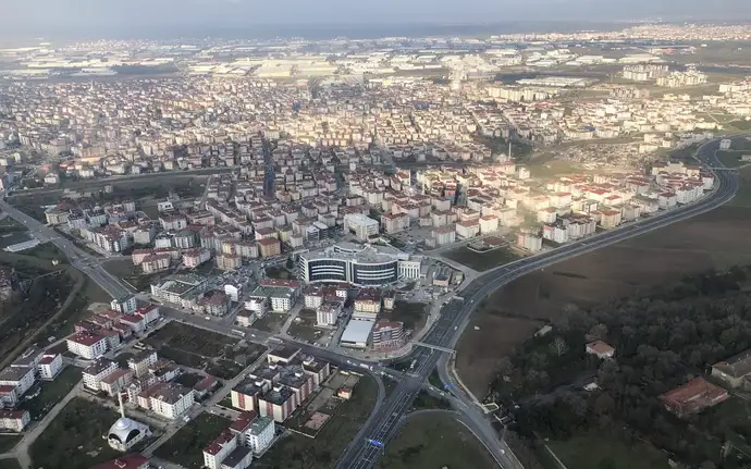 Aerial view of Çerkezköy city with rows of mid‑rise apartment buildings, wide roads, and a large modern complex at the center, surrounded by open fields and patches of forest under a partly cloudy sky