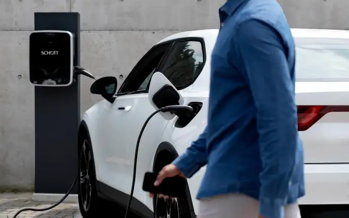 A man walks past a white electric car charging at a station.