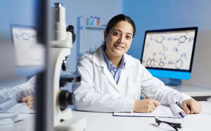 Smiling scientist at a lab desk with a microscope, chemical formulas on a screen behind her.