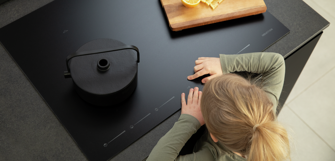 Top view of black glass cooktop with pot, cutting board with lemon slices, and person nearby