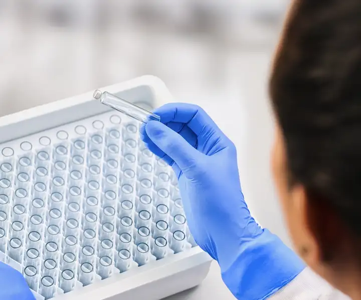 A laboratory worker in blue gloves holding a small glass vial, with a tray of vials in the background.