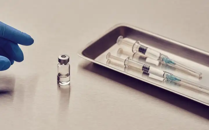 Gloved hand reaching toward a small vial beside three syringes on a metal tray